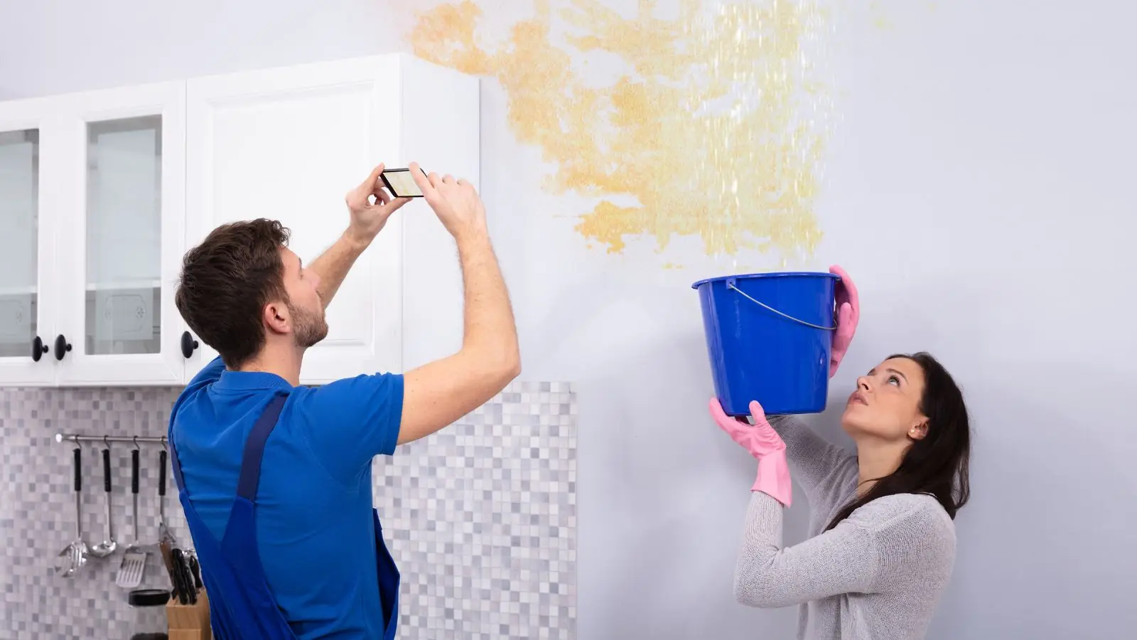 Man photographing a water-stained wall while a woman holds a bucket to catch a leak
