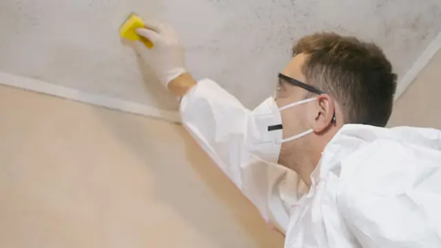 Person in a mask, goggles and protective suit wiping a stained ceiling with a sponge